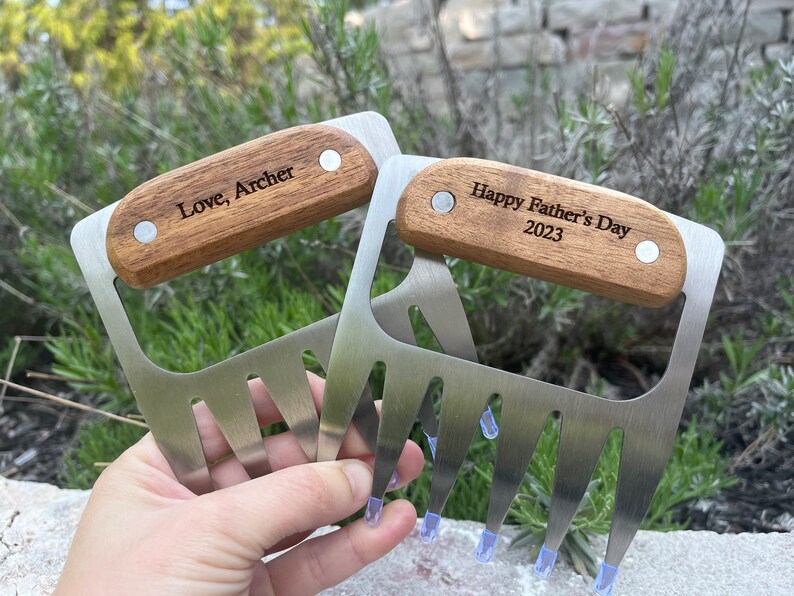 May include: Two stainless steel meat shredder claws with wooden handles. One claw is engraved with "Love, Archer", the other with "Happy Father's Day 2023". The claws have blue plastic tips.
