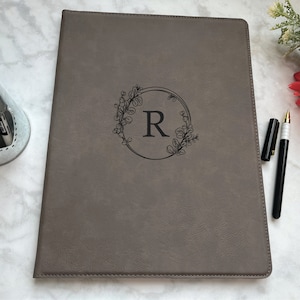 May include: A gray leather-bound journal with a floral wreath design and the letter "R" in the center. The journal is on a white marble surface with two black pens.