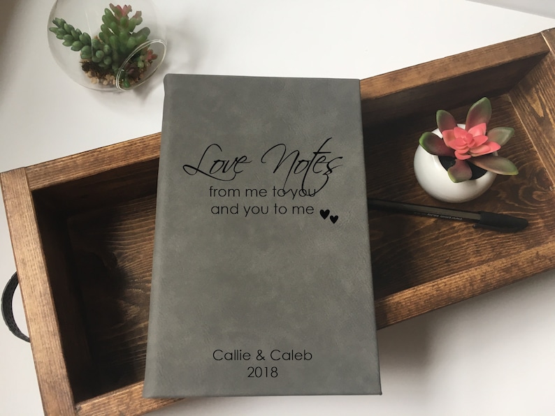 Op de afbeelding: Een grijs lederen gebonden dagboek met de tekst "Love Notes from me to you and you to me" en twee kleine hartjes. Het dagboek is gepersonaliseerd met de namen "Callie & Caleb" en het jaar "2018".