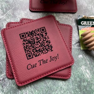 May include: Burgundy-colored square coasters with a QR code and the text "Cue The Joy!". The coasters are stacked, with a tea bag and wooden honey dipper in the background. The coasters are made of a textured material.