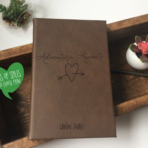 Puede incluir: Un diario de cuero marr&oacute;n con el texto "Adventure Awaits" y una flecha que atraviesa un coraz&oacute;n. El diario est&aacute; personalizado con el nombre "Lanie Mae".
