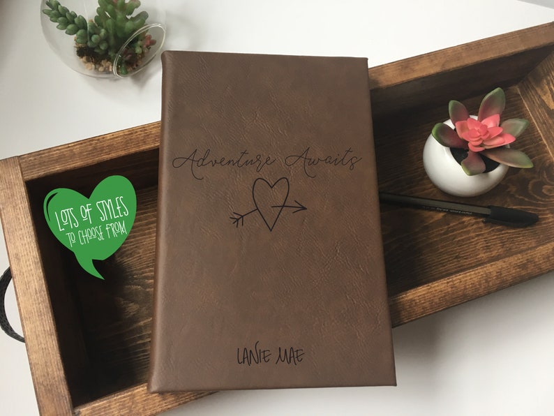 Puede incluir: Un diario de cuero marr&oacute;n con el texto "Adventure Awaits" y una flecha que atraviesa un coraz&oacute;n. El diario est&aacute; personalizado con el nombre "Lanie Mae".