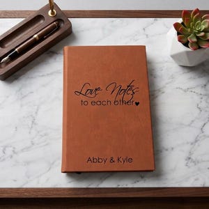 May include: A tan leather-bound journal featuring the black printed text Love Notes to each other with a small heart icon and the names Abby & Kyle at the bottom. The journal is positioned on a white marble tray with a wooden frame and pen holder.