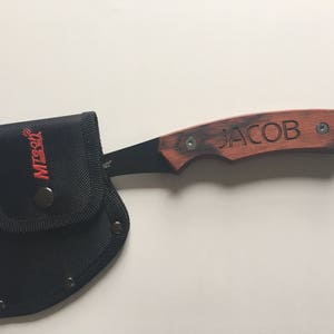 May include: A small black and brown axe with a wooden handle. The handle is engraved with the name "JACOB". The axe comes with a black nylon sheath.
