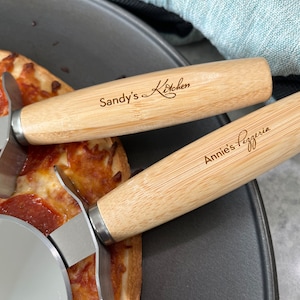 May include: Two wooden pizza cutters with metal blades. The top cutter has the text "Sandy's Kitchen" engraved on the handle. The bottom cutter has the text "Annie's Pizzeria" engraved on the handle.