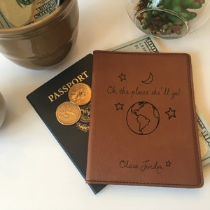 May include: A black passport with gold lettering and coins next to a brown leather passport cover. The cover is engraved with the phrase "Oh the places she'll go!" and a world map, stars, and moon design. The name "Olivia Jordyn" is also engraved.