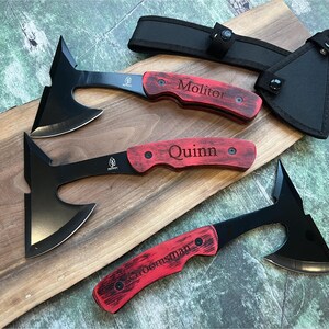 May include: Three black hatchet-style throwing knives with red wooden handles. The handles are engraved with the names "Molitor", "Quinn", and "Sroensman".