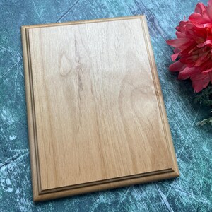 May include: A rectangular wooden plaque with a light brown finish and a decorative border. The plaque is displayed on a green and blue surface, with a pink flower and greenery in the background.