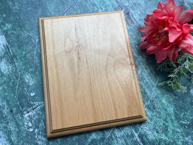 May include: A rectangular wooden plaque with a light brown finish and a decorative border. The plaque is displayed on a green and blue surface, with a pink flower and greenery in the background.