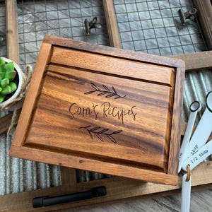 May include: A rectangular wooden recipe box with a dark brown grain, featuring the engraved text "Sara's Recipes" centred between two leaf illustrations. The lid has a recessed panel design, and the box is crafted from polished wood.