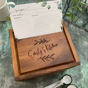 May include: A rectangular wooden recipe box with a dark brown finish. The lid is engraved with the text "Emily's Kitchen" flanked by two leaf sprigs. A white recipe card with green eucalyptus accents is tucked into the box, displaying the headings "Recipe," "From the home of," "Ingredients," and "Directions."