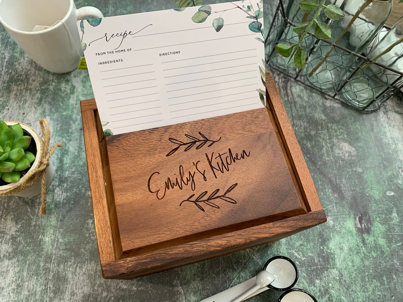 May include: A rectangular wooden recipe box with a dark brown finish. The lid is engraved with the text "Emily's Kitchen" flanked by two leaf sprigs. A white recipe card with green eucalyptus accents is tucked into the box, displaying the headings "Recipe," "From the home of," "Ingredients," and "Directions."