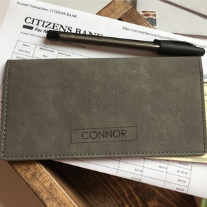 Puede incluir: Una funda rectangular gris para chequera con superficie texturizada y el nombre CONNOR grabado en un marco rectangular negro en la parte frontal. La funda presenta costuras visibles a lo largo de los bordes y se coloca sobre un documento bancario.