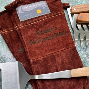 K&ouml;nnte beinhalten: Ein Set Grillwerkzeuge, darunter ein Spatel, Fleischkrallen und hitzebest&auml;ndige Handschuhe. Die Handschuhe sind braun mit dem Text "Daddy's Backyard BBQ" und einem Etikett mit der Aufschrift "Grab life by the grills". Spatel und Fleischkrallen haben Holzgriffe.