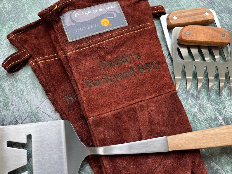 K&ouml;nnte beinhalten: Ein Set Grillwerkzeuge, darunter ein Spatel, Fleischkrallen und hitzebest&auml;ndige Handschuhe. Die Handschuhe sind braun mit dem Text "Daddy's Backyard BBQ" und einem Etikett mit der Aufschrift "Grab life by the grills". Spatel und Fleischkrallen haben Holzgriffe.