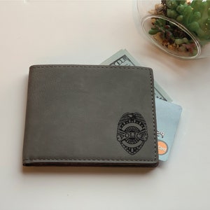 May include: A grey bi-fold wallet featuring a laser-engraved police badge emblem on the front. The wallet contains a visible paper currency bill and a credit card with a visible Mastercard logo peeking out from the top edge of the wallet.