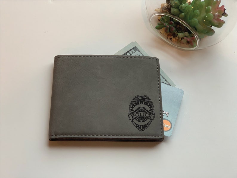 May include: A grey bi-fold wallet featuring a laser-engraved police badge emblem on the front. The wallet contains a visible paper currency bill and a credit card with a visible Mastercard logo peeking out from the top edge of the wallet.