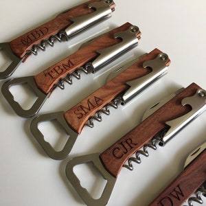 May include: Five personalized wooden bottle openers with corkscrews. Each opener has a different name engraved on the handle: MBD, TBM, SMA, CJR, and DW.