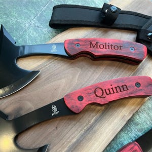 May include: Two black tactical tomahawks with red wooden handles. The handles are engraved with the names "Molitor" and "Quinn".