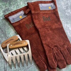 May include: A set of brown suede grilling gloves with the word "Dad" printed on them, along with two stainless steel meat shredder claws with wooden handles. The gloves have a patch that says "Grab life by the grill".
