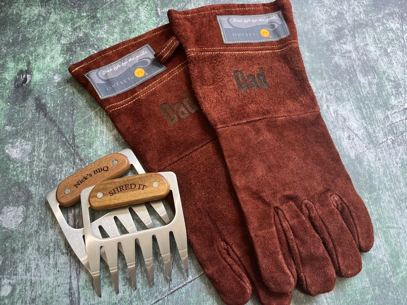 May include: A set of brown suede grilling gloves with the word "Dad" printed on them, along with two stainless steel meat shredder claws with wooden handles. The gloves have a patch that says "Grab life by the grill".