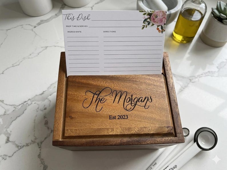 May include: A rectangular wooden recipe box with a lid engraved with the text The Morgans Est 2023. A white recipe card with floral accents and the text This Dish, Prep Time & Serves, Ingredients, and Directions is inserted into the box lid slot.