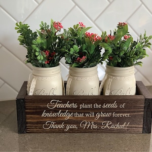 May include: A wooden box with three mason jars filled with greenery and red flowers. The box has the text "Teachers plant the seeds of knowledge that will grow forever. Thank you, Mrs. Rachul!" engraved on the front.