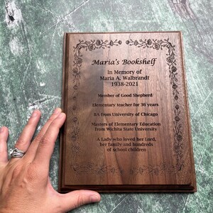 May include: A wooden plaque with the text "Maria's Bookshelf" and an inscription honoring Maria A. Walbrandt, including her birth and death years, educational background, and a tribute. The plaque has a floral border.