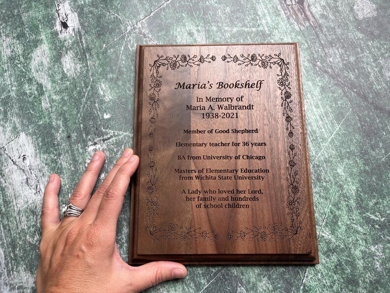 May include: A wooden plaque with the text "Maria's Bookshelf" and an inscription honoring Maria A. Walbrandt, including her birth and death years, educational background, and a tribute. The plaque has a floral border.