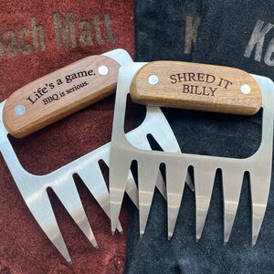 May include: Two stainless steel meat shredder claws with wooden handles. One handle reads "Life's a game. BBQ is serious." and the other reads "SHRED IT BILLY." The claws are on brown and black surfaces.
