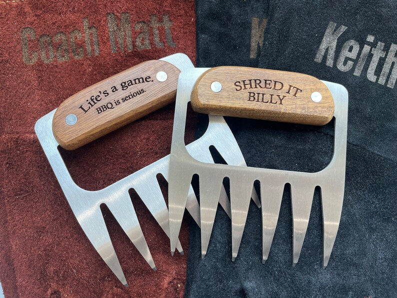 May include: Two stainless steel meat shredder claws with wooden handles. One handle reads "Life's a game. BBQ is serious." and the other reads "SHRED IT BILLY." The claws are on brown and black surfaces.