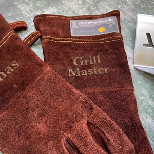K&ouml;nnte beinhalten: Ein Paar braune Wildleder-Grillhandschuhe mit dem Namen "Jonas" und dem Aufdruck "Grill Master". Ein Edelstahl-Pfannenwender liegt neben den Handschuhen. Die Handschuhe haben ein Etikett mit der Aufschrift "Outset".