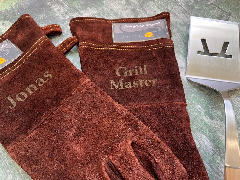 K&ouml;nnte beinhalten: Ein Paar braune Wildleder-Grillhandschuhe mit dem Namen "Jonas" und dem Aufdruck "Grill Master". Ein Edelstahl-Pfannenwender liegt neben den Handschuhen. Die Handschuhe haben ein Etikett mit der Aufschrift "Outset".