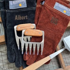 K&ouml;nnte beinhalten: Schwarze und braune Leder-Grillhandschuhe mit dem Namen "Albert" und den Initialen "TJS" aufgestickt. Zwei Fleischkrallen aus Edelstahl mit Holzgriffen und ein Metallspatel sind ebenfalls zu sehen. Die Handschuhe haben ein "Outset"-Etikett.