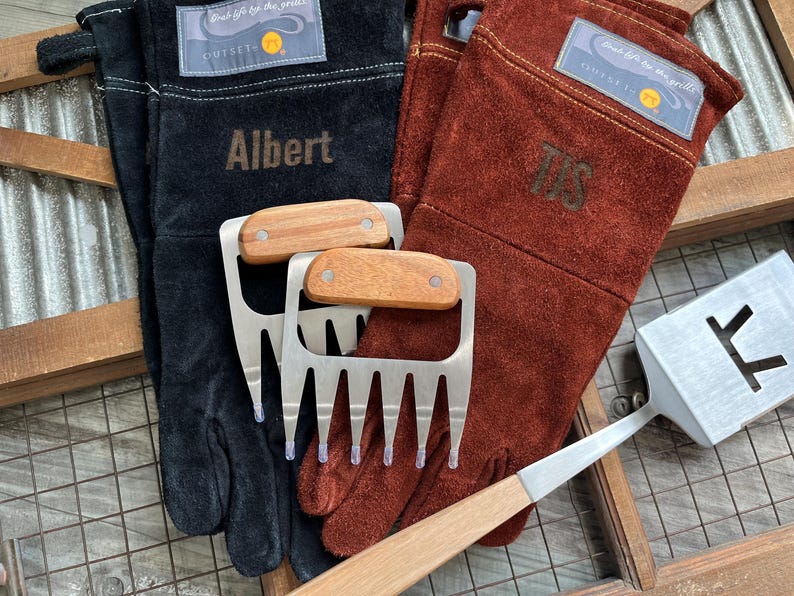 K&ouml;nnte beinhalten: Schwarze und braune Leder-Grillhandschuhe mit dem Namen "Albert" und den Initialen "TJS" aufgestickt. Zwei Fleischkrallen aus Edelstahl mit Holzgriffen und ein Metallspatel sind ebenfalls zu sehen. Die Handschuhe haben ein "Outset"-Etikett.