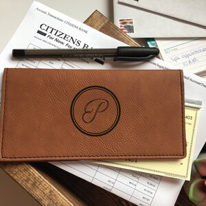 May include: A rectangular tan faux leather checkbook cover featuring a dark brown circular emblem with a stylized letter P in the center. The cover has visible black stitching along the edges and is positioned over financial documents and a black pen.