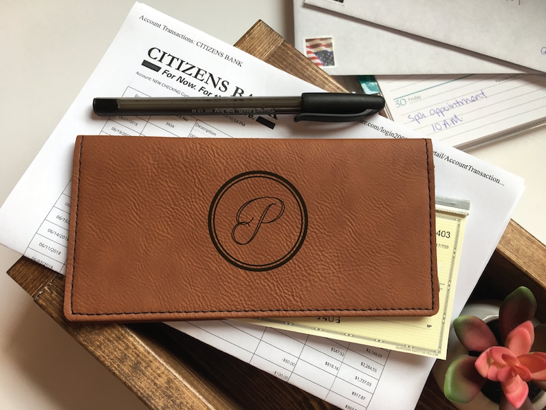 May include: A rectangular tan faux leather checkbook cover featuring a dark brown circular emblem with a stylized letter P in the center. The cover has visible black stitching along the edges and is positioned over financial documents and a black pen.