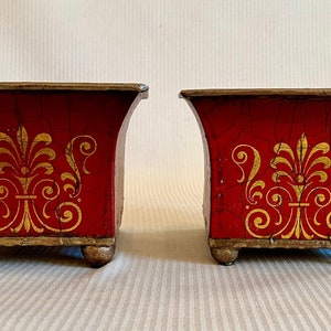 Può includere: Due vasi da fiori in metallo rosso con disegni dipinti in oro. I vasi hanno una base quadrata e una parte superiore svasata. Ogni vaso ha quattro piccoli piedini.
