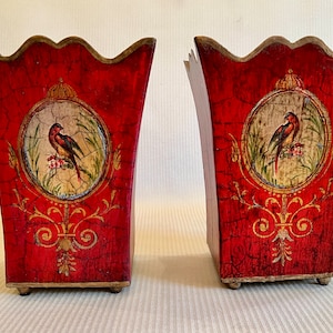 Puede incluir: Un par de jarrones decorativos de madera roja con un acabado desgastado. Cada jarrón presenta un diseño pintado en oro y un motivo circular con un pájaro y detalles florales.