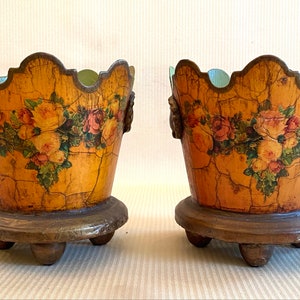 Könnte beinhalten: Zwei dekorative Pflanzentopf aus Holz mit einer abgenutzten Oberfläche. Die Töpfe sind mit einem floralen Design in Gelb-, Rosa- und Grüntönen bemalt. Jeder Topf hat einen gewellten Rand und steht auf einem Holzsockel mit drei Beinen.