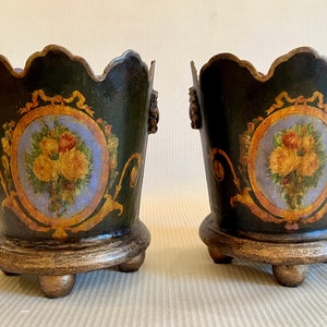 Puede incluir: Un par de macetas de metal pintadas en negro y dorado con diseños florales. Cada maceta tiene un borde festoneado y se asienta sobre tres patas.