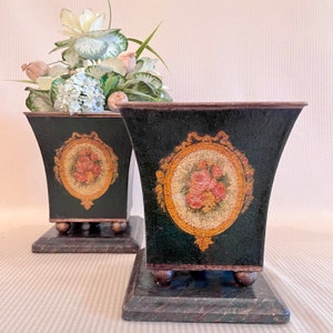 May include: Two black metal planters with gold accents and floral designs. The planters have a square base and a flared top. They are decorated with a gold oval frame with a floral design inside. The planters are sitting on a striped surface.