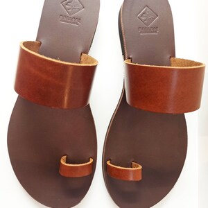 May include: A pair of brown leather sandals with a toe loop and a wide strap across the top. The sandals have a dark brown sole and a lighter brown strap. The brand name is stamped on the heel.
