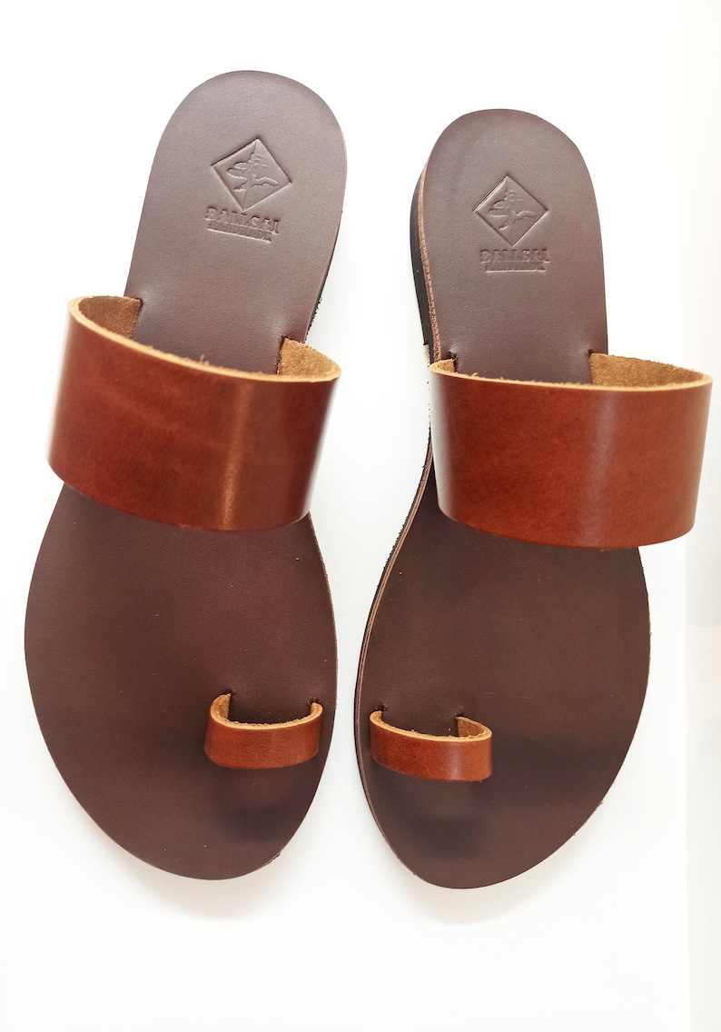 May include: A pair of brown leather sandals with a toe loop and a wide strap across the top. The sandals have a dark brown sole and a lighter brown strap. The brand name is stamped on the heel.