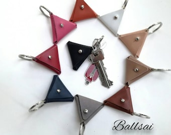 May include: A collection of ten leather triangle keychains in various colors, including red, pink, white, brown, black, and gray. Each keychain features a silver ring and a small metal button. The keychains are arranged in a circle around a set of keys with a red and silver charm.