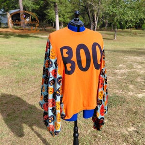 May include: Orange long-sleeved shirt with the word "BOO" in black block letters. The sleeves have a light blue background with a pattern of orange and black jack-o'-lanterns. A festive Halloween-themed shirt.