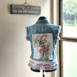 May include: A light blue denim vest with a tapestry angel design on the back. The vest has pink fringe along the edges and bottom. A sign above reads "All things are possible for one who believes!"