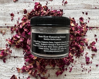 May include: A white jar with a black lid and label that reads "Rose Glow Cleansing Grains Gentle Exfoliation White Clay, Organic Oats, Organic Almonds, Roses, Organic Orange & Rose Essential Oil Scoop a little, mix with water, gently rub over rinse clean. Topical use only. thewildapothecary.com"