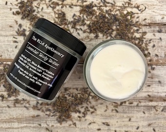 May include: A glass jar of lavender body butter with a black label that reads "The Wild Apothecary Lavender Body Butter". The ingredients are listed on the label: Apricot Oil, Shea Butter*Golden Ajoba Oil, Coconut Oil, Lavender Essential Oil, Arrowroot Powder. The jar is sitting on a wooden surface with dried lavender flowers scattered around it.