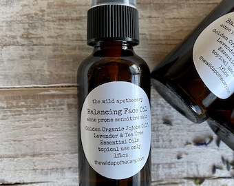 May include: A brown glass bottle with a black pump top and a white label that reads "the wild apothecary Balancing Face Oil acne prone sensitive skin Golden Organic Jojoba Oil, Lavender & Tea Tree Essential Oils topical use only 1 fl oz thewildapothecary.com"